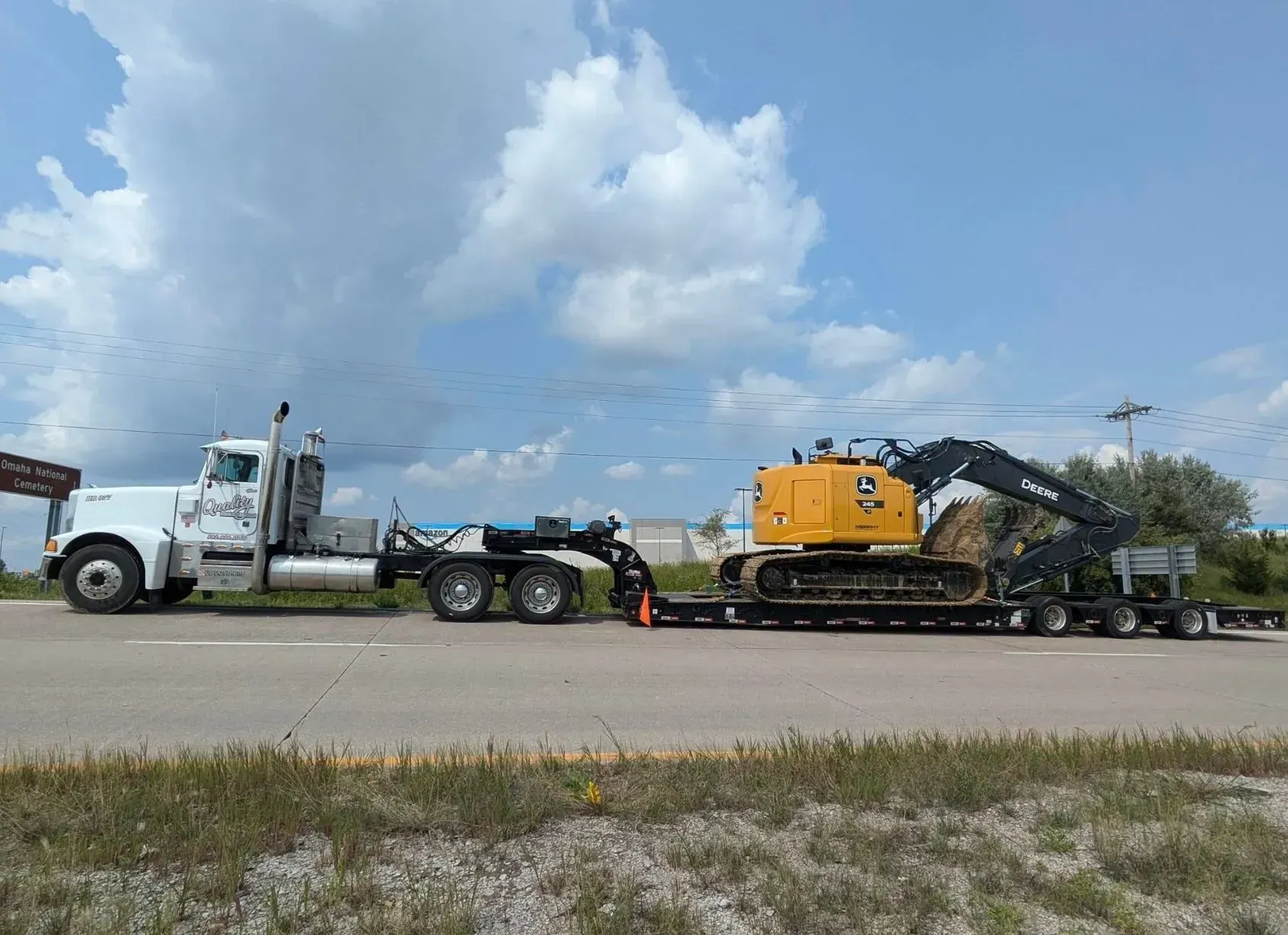 White semi-truck hauling a yellow excavator on a flatbed trailer on a road under a blue sky.