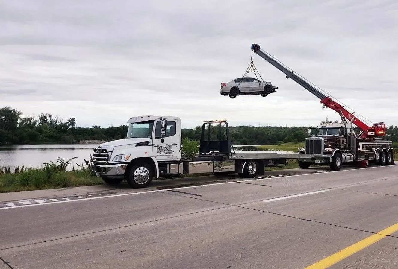 Tow trucks lifting a car from roadside. Cloudy sky, water, and trees in background.