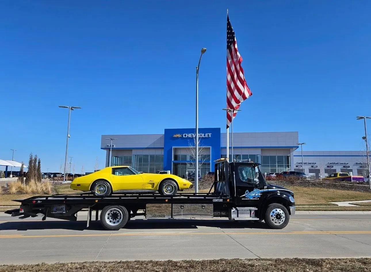 Yellow Corvette being transported on a flatbed tow truck in front of a Chevrolet dealership. American flag in the background.