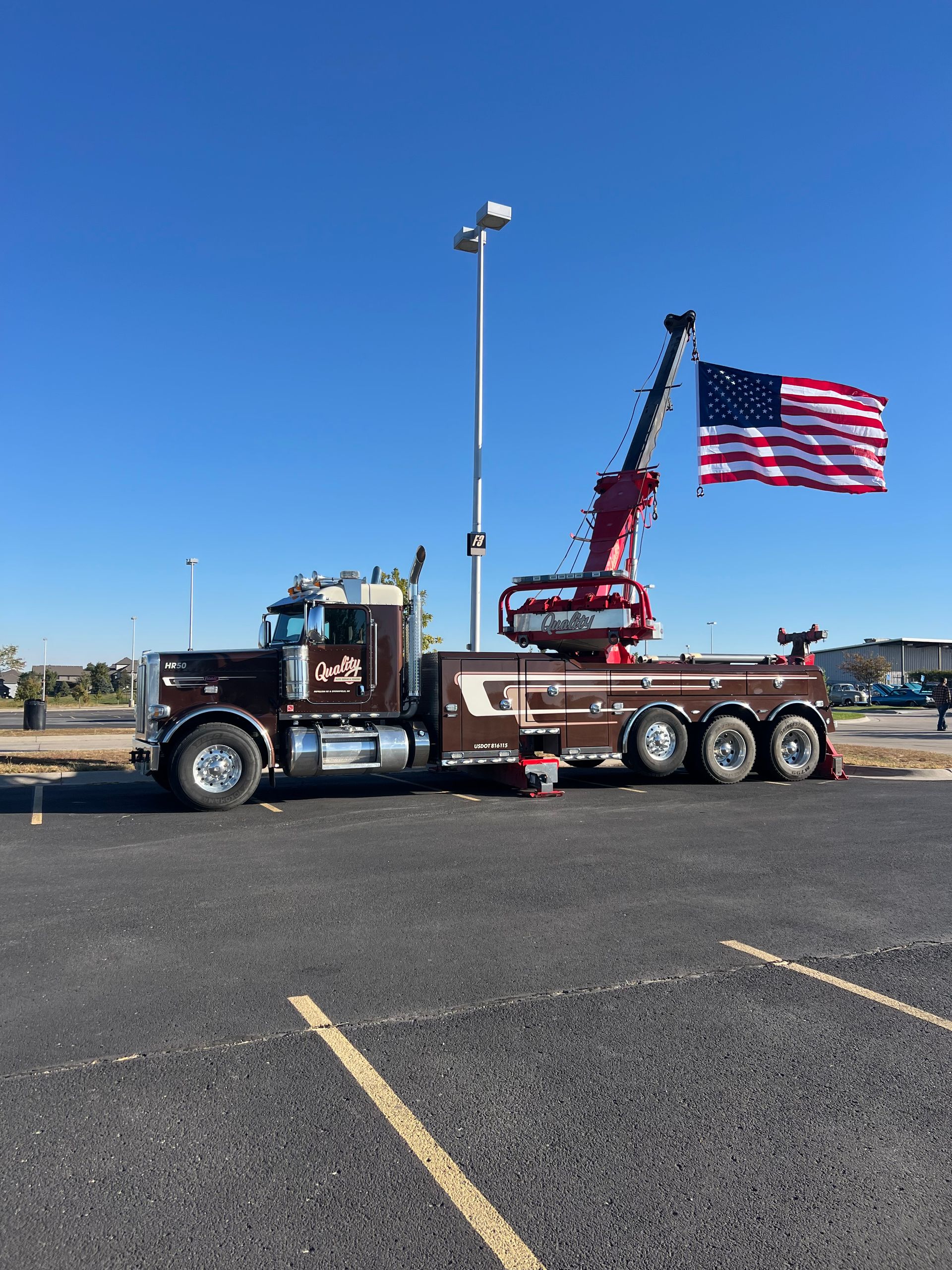 Dark brown tow truck with an American flag attached to a crane in a parking lot.