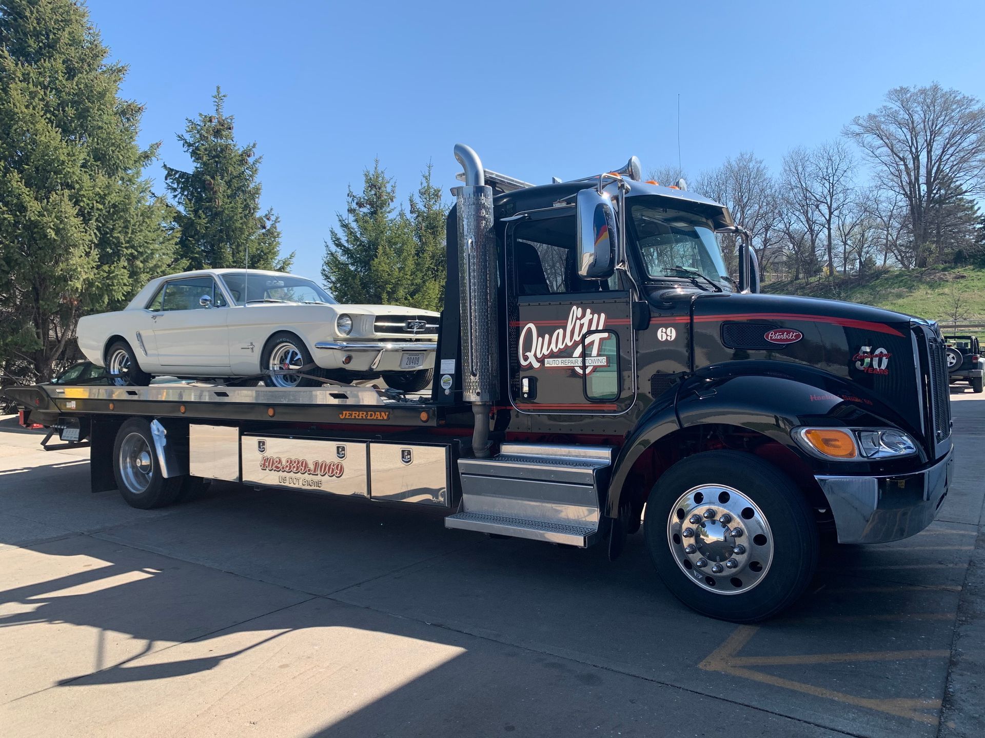 Black tow truck carrying a white vintage Ford Mustang on a sunny day.