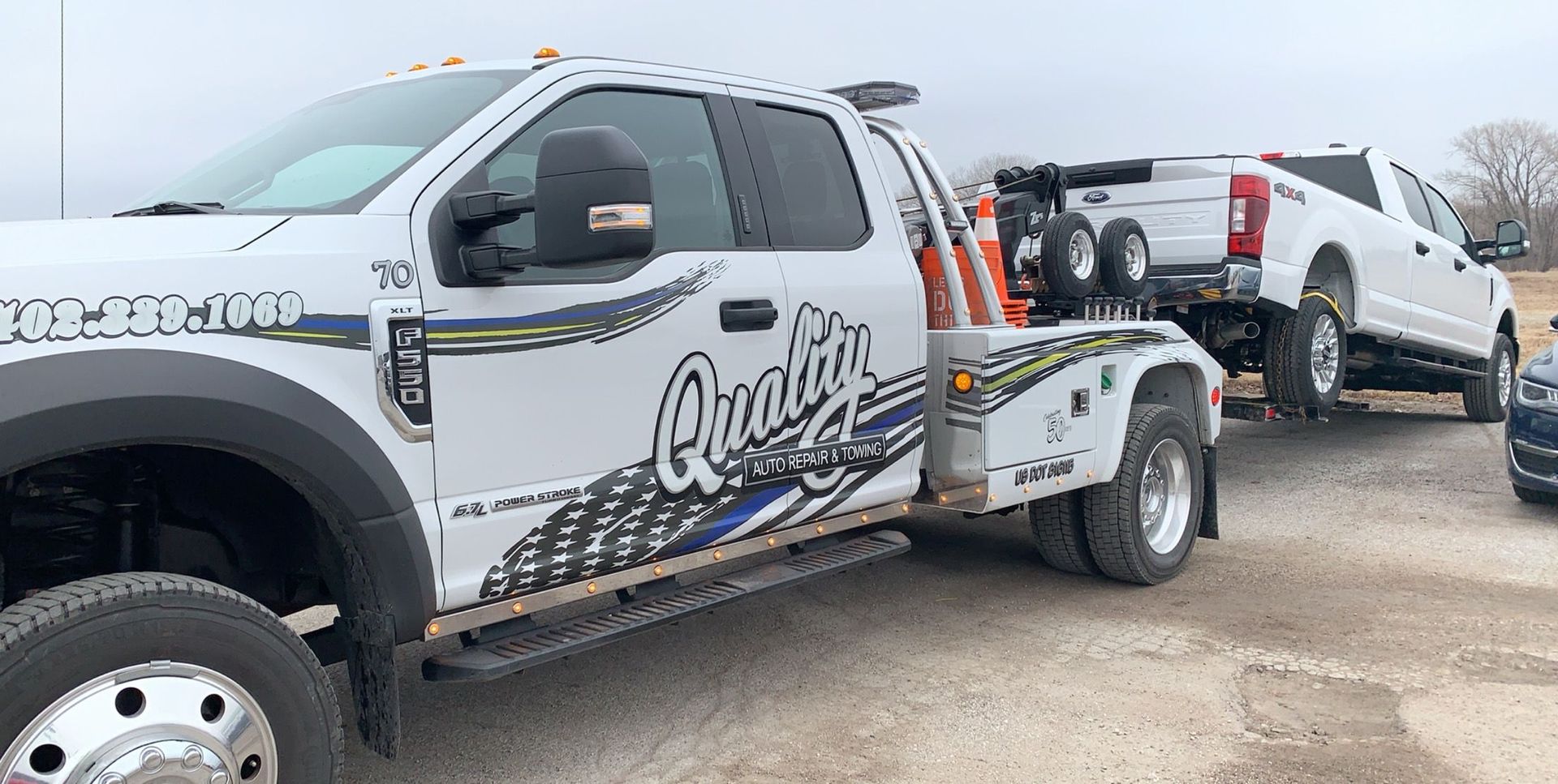 A white tow truck towing another white truck on a dirt road, cloudy sky.