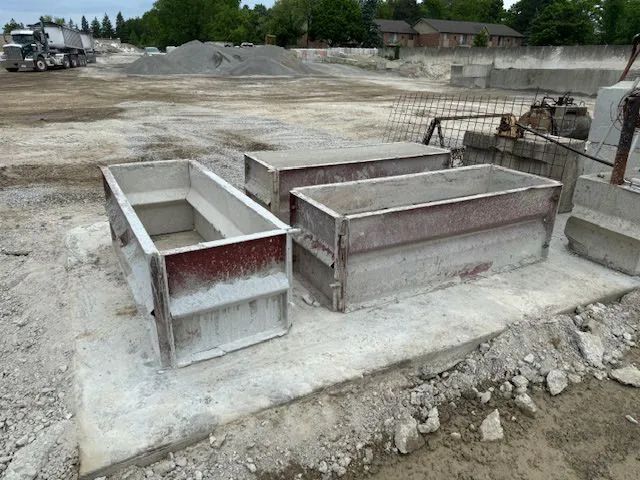 Three rectangular concrete forms on a construction site, with gray rock piles and a truck in the background.