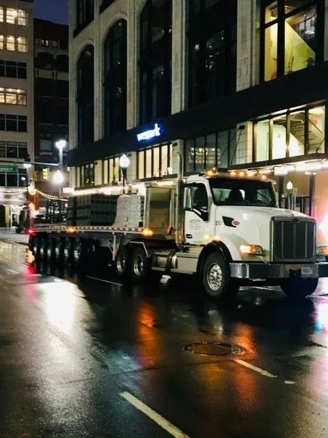 Flatbed truck parked on wet city street at night. Buildings with lit windows in background.