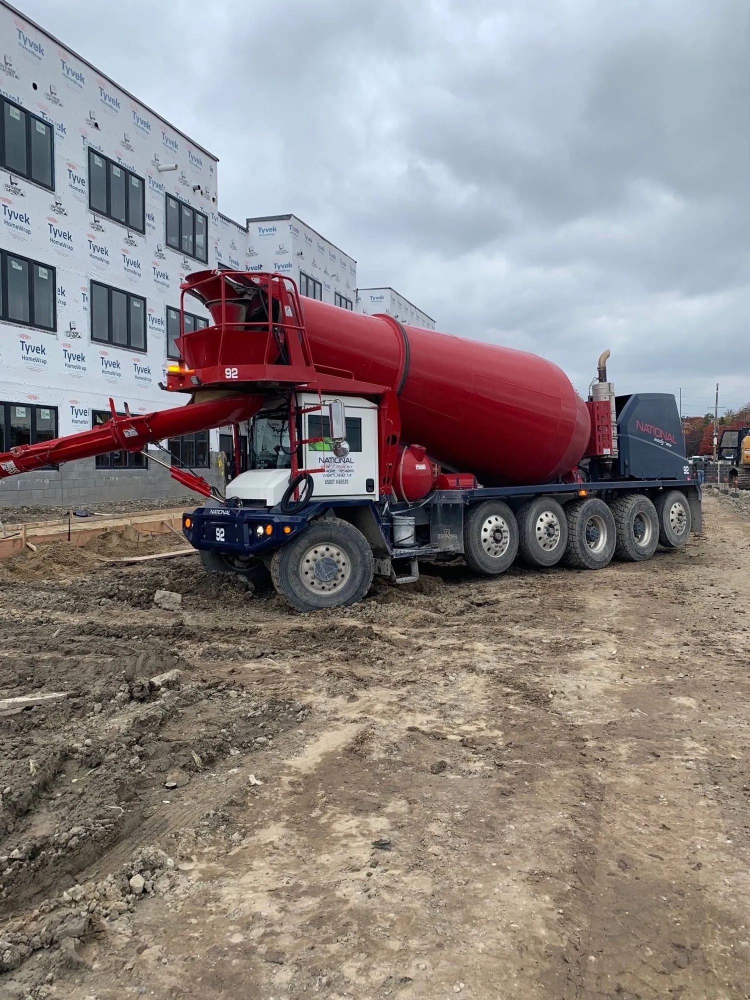 Red concrete mixer truck on a muddy construction site, with extended boom. Building in background.
