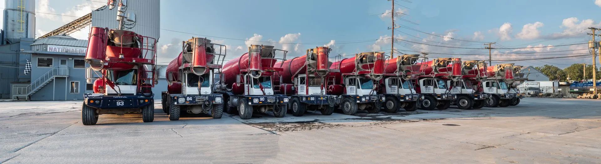 A row of red and white cement trucks parked outside a building on a sunny day.