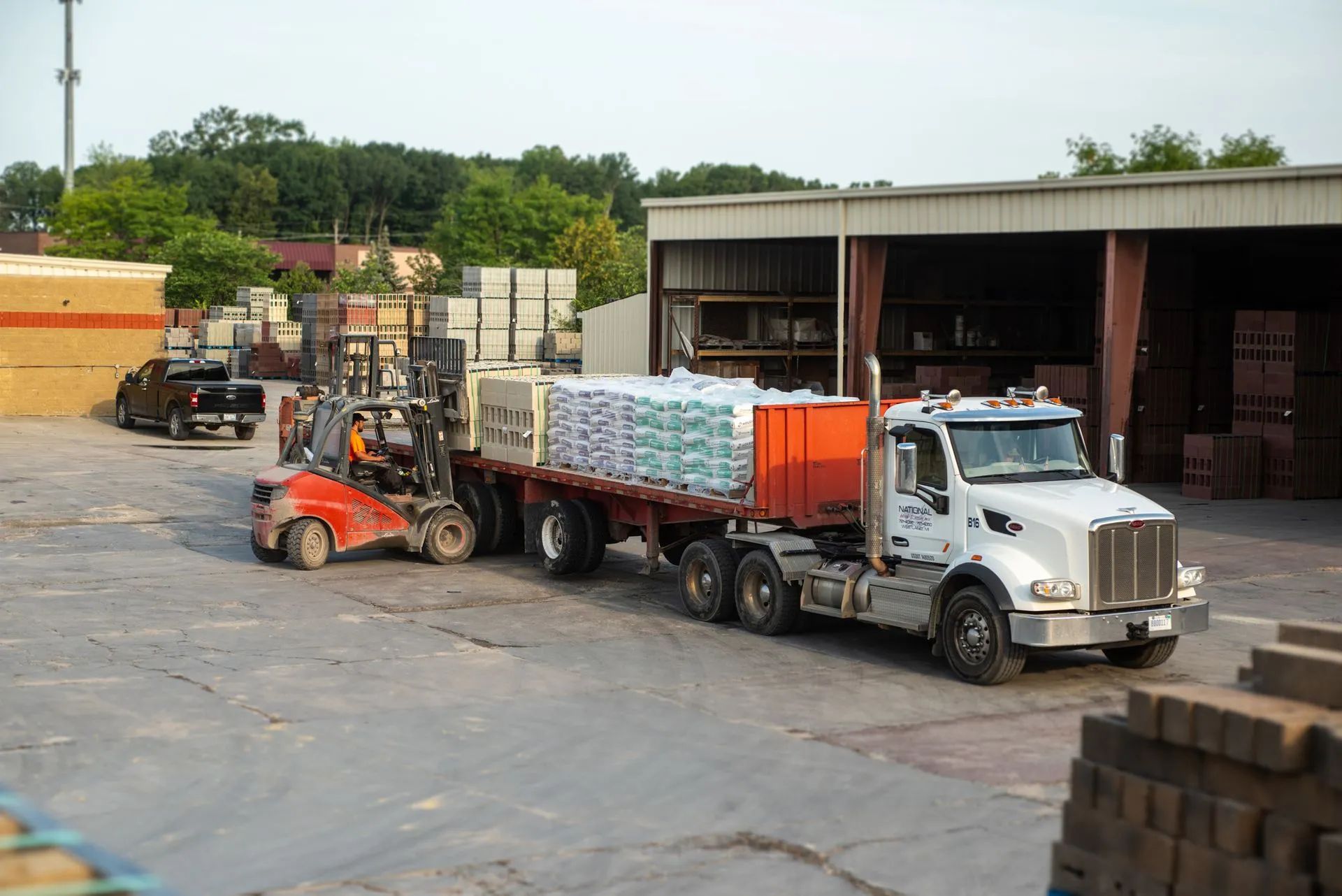 Forklift loading pallets of materials onto a semi-truck trailer in a warehouse yard.