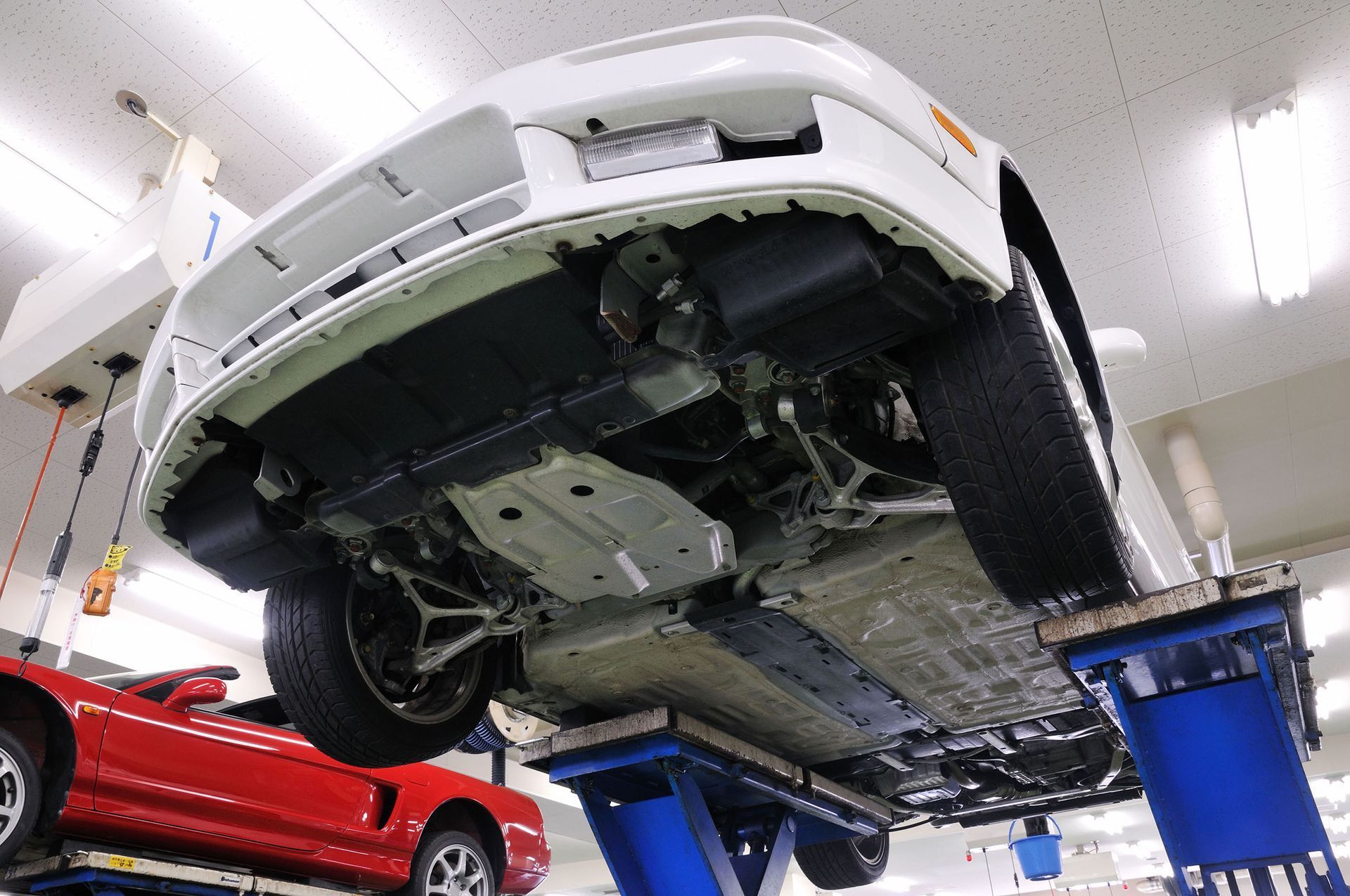 White car on a lift in a garage, showing undercarriage. A red car is partially visible.