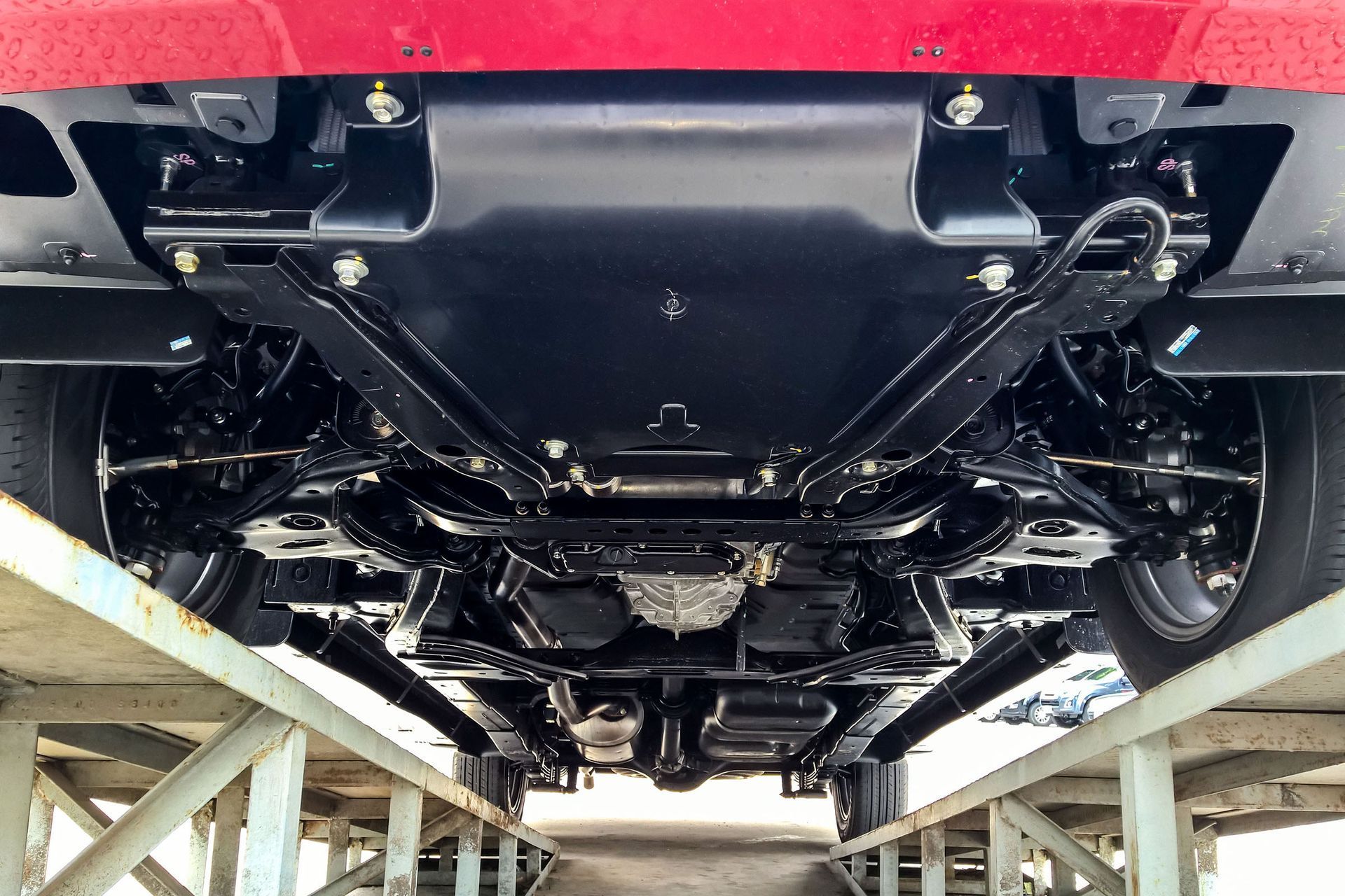 Undercarriage of a vehicle, viewed from below, painted black and supported on a platform, showing suspension, engine components, and wheels.
