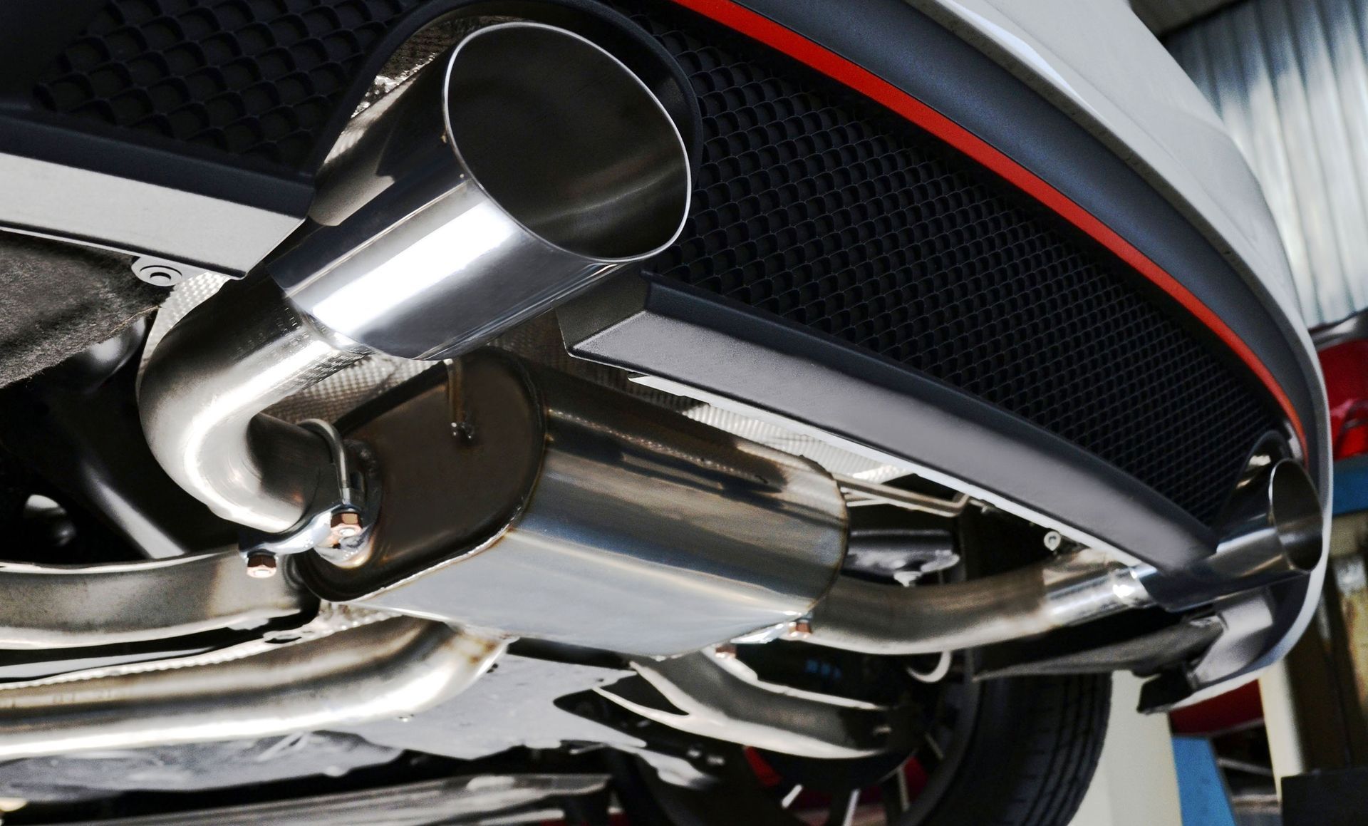 Shiny chrome exhaust system under a white car with a black mesh grille and red trim.