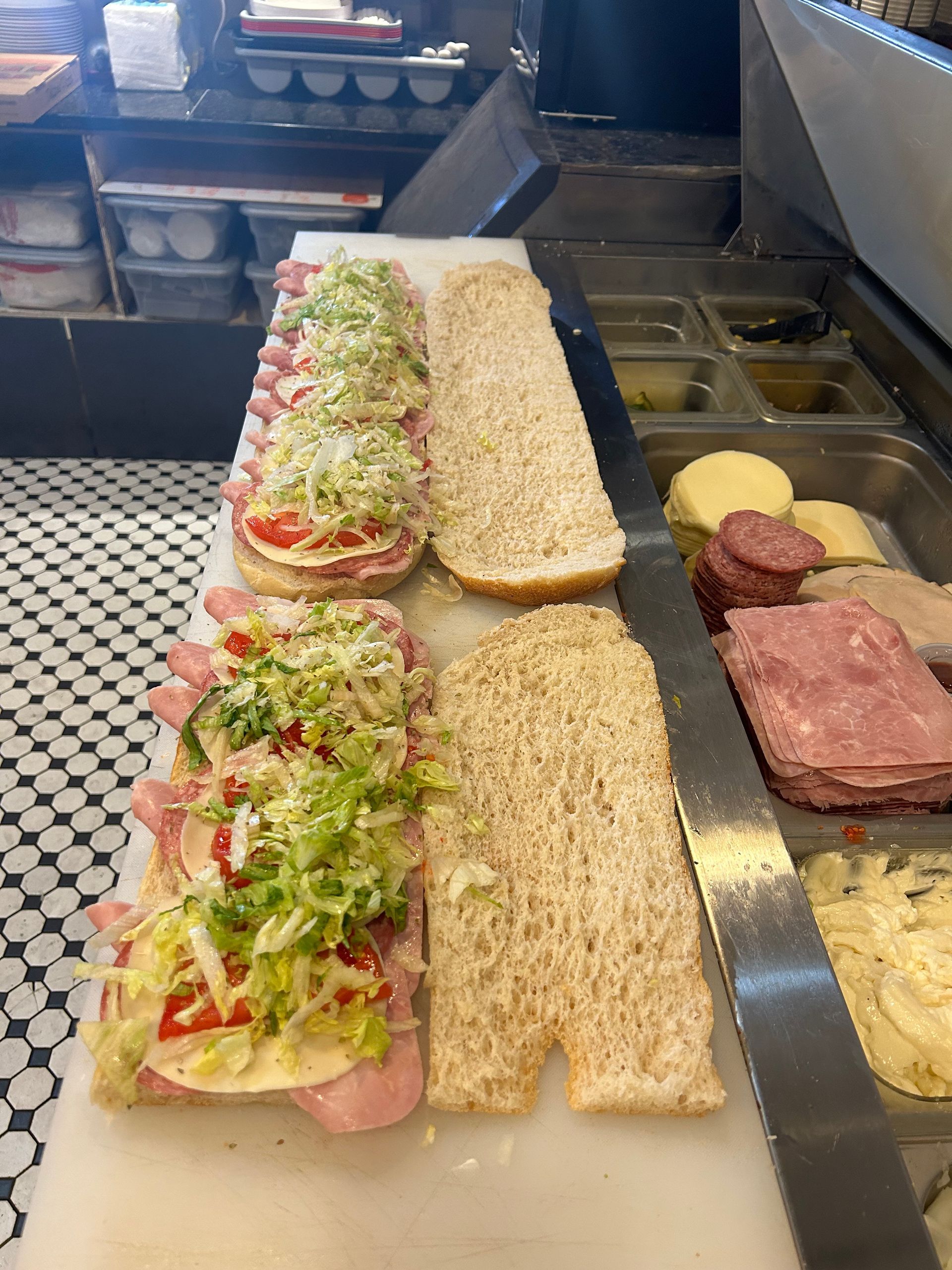 A sub sandwich is being prepared on a cutting board in a kitchen.