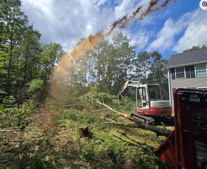 Excavator clearing brush on a wooded hillside near a house, with debris spraying into the sky