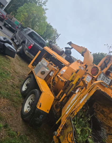 Orange wood chipper hitched to a truck, angled on a grassy roadside with parked vehicles nearby