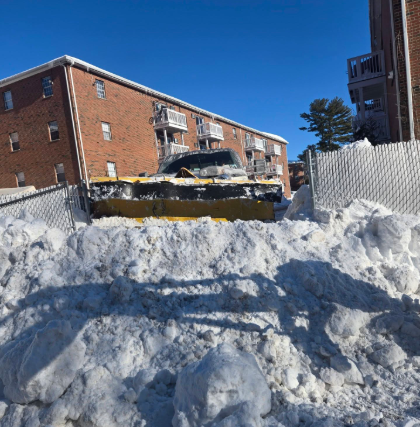 Snow-covered parking lot with a yellow snowplow clearing deep snow beside brick apartment buildings.