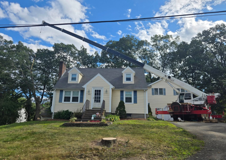 House with steep roof and chimney being repaired by a red crane in a wooded yard under blue sky