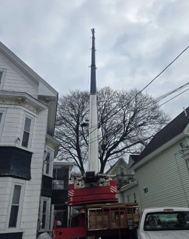 Crane lifting a large white section over a red truck between houses on a cloudy street