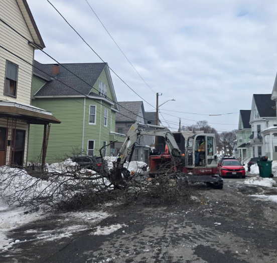 Snowy residential street with a backhoe clearing mud and debris beside houses
