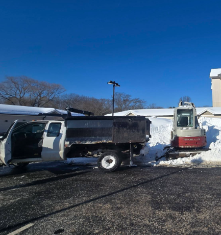 White dump truck beside a red skid steer in a snowy parking lot under a clear blue sky