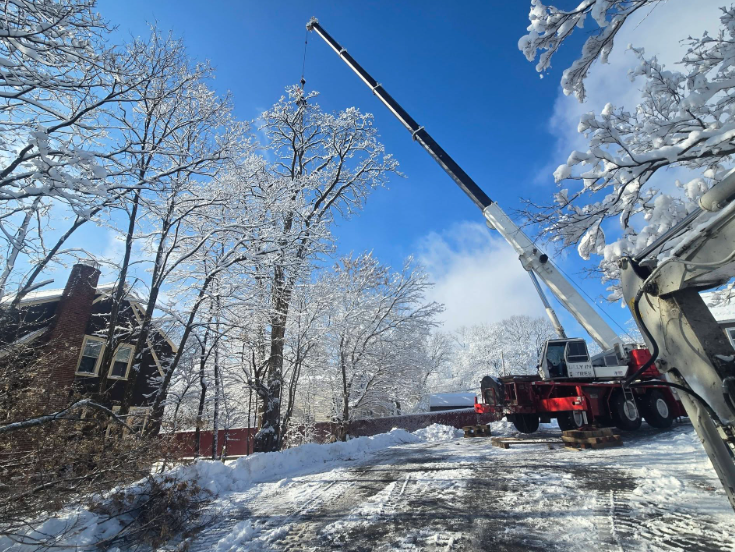 Snow-covered street with a crane truck clearing ice beneath frosty trees and a blue sky