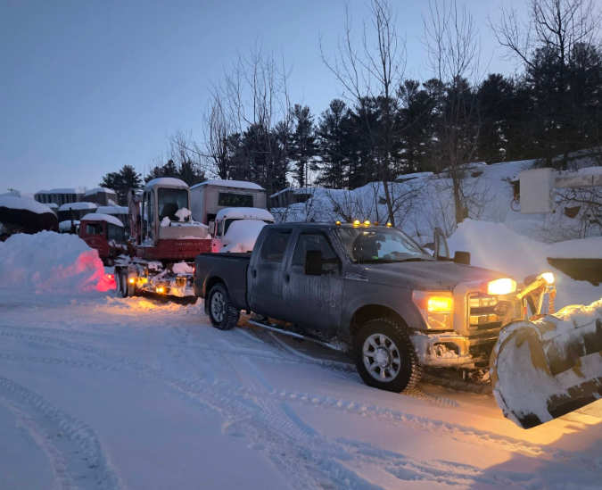 Two snowplow trucks clearing a snowy road at dusk, headlights glowing in a winter landscape