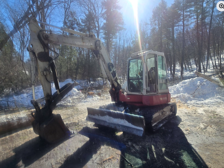 Red mini excavator with bucket in a snowy wooded area, bright sun overhead.