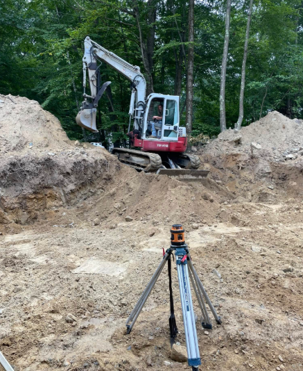 Excavator digging earth in a wooded construction site, with a tripod survey instrument in the foreground