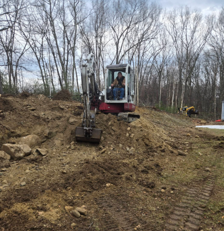 Small excavator on a dirt mound in a wooded clearing, with a worker inside the cab.