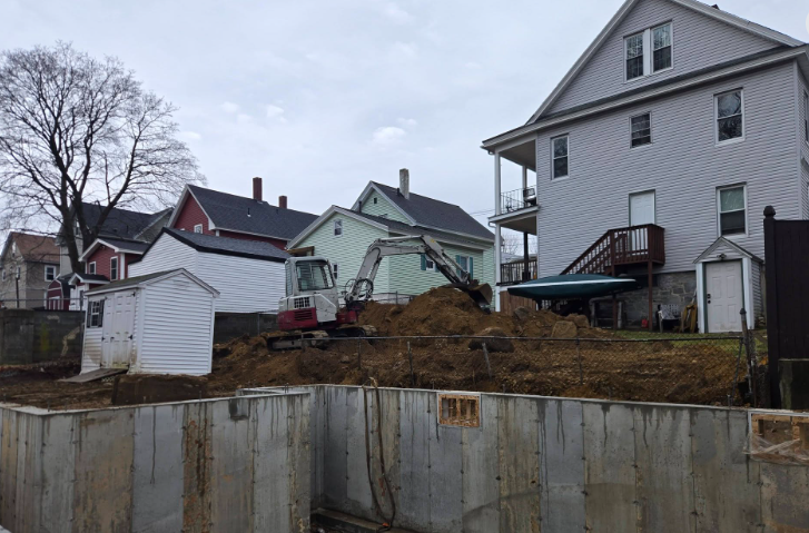 Excavator on a muddy lot beside houses with a retaining wall and bare trees under a gray sky