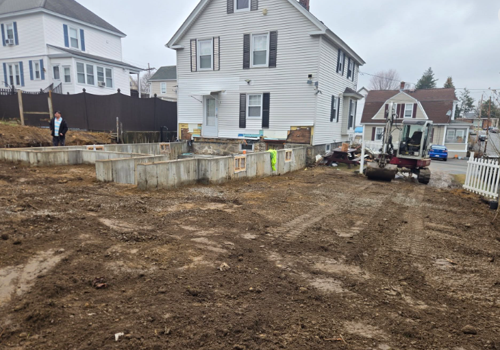 Construction site with excavated dirt, concrete foundation, and a small utility vehicle beside houses