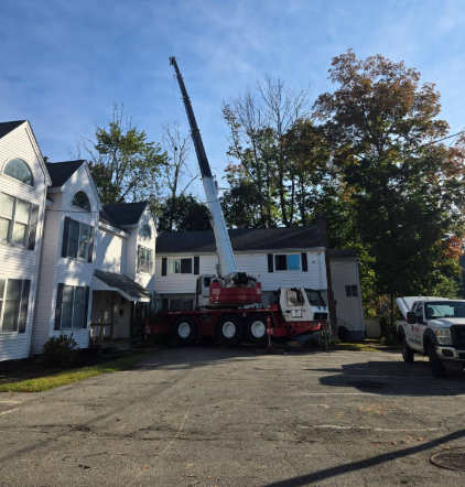 Crane truck parked outside white houses on a sunny street, with a raised boom and a parked utility vehicle nearby
