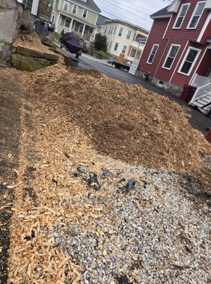 Wood chip-covered sloped lot beside houses, with gravel and a person in the background