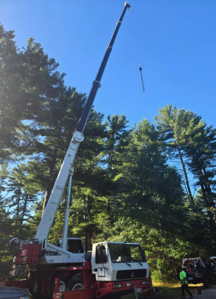 Crane lifting a long cable near tall pine trees under a clear blue sky