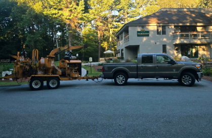 Dark pickup truck towing a yellow wood chipper trailer on a road near a house and trees at dusk