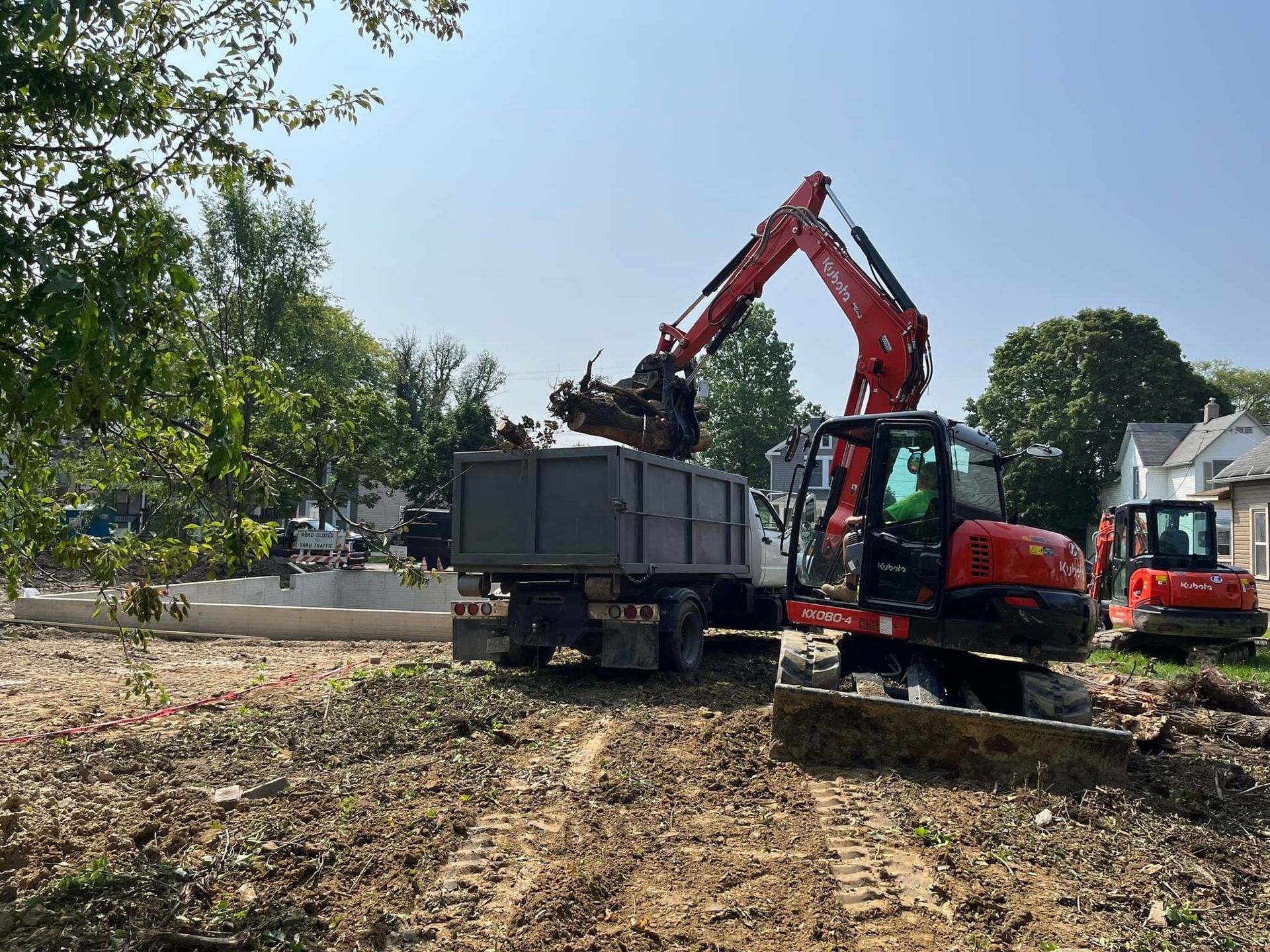 A red excavator is loading a dumpster in a dirt field.