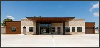 A modern, one-story commercial building with light stone, wood paneling, a dark overhang, and a large central glass entrance.