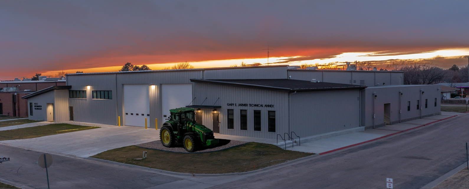 A green John Deere tractor parked outside a gray building with windows; sunset in the background.