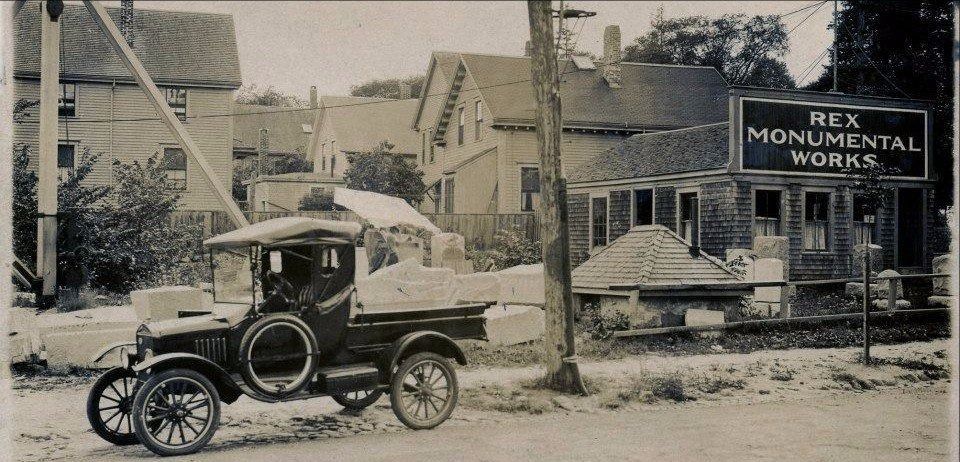 An old car is parked in front of a building that says Rex Monumental Works