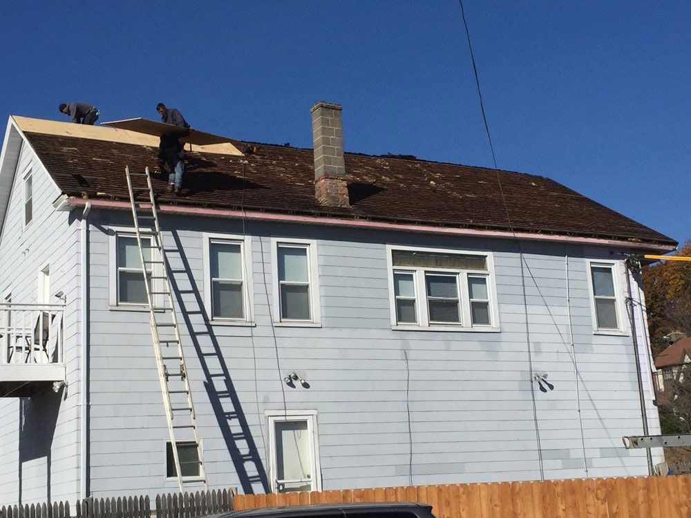 Two workers install new roof sheeting on the sloped roof of a light blue house with a ladder leaning against the side.