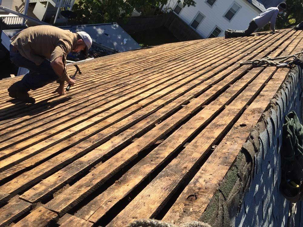 Two people work on a sloped roof, nailing wood slats to the roof deck on a sunny day.
