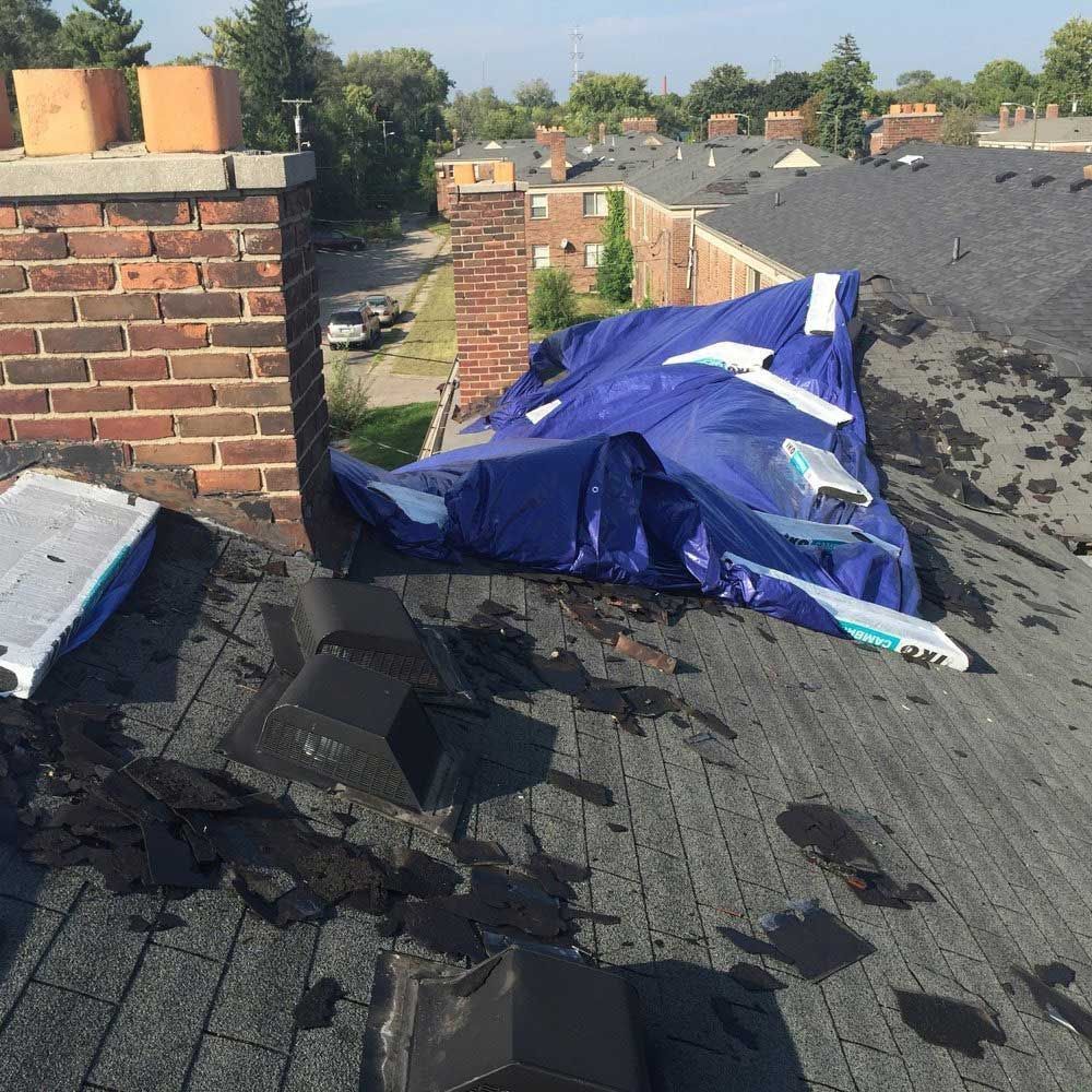 A damaged residential roof with missing shingles, black vents, a brick chimney, and a large blue tarp covering a section.