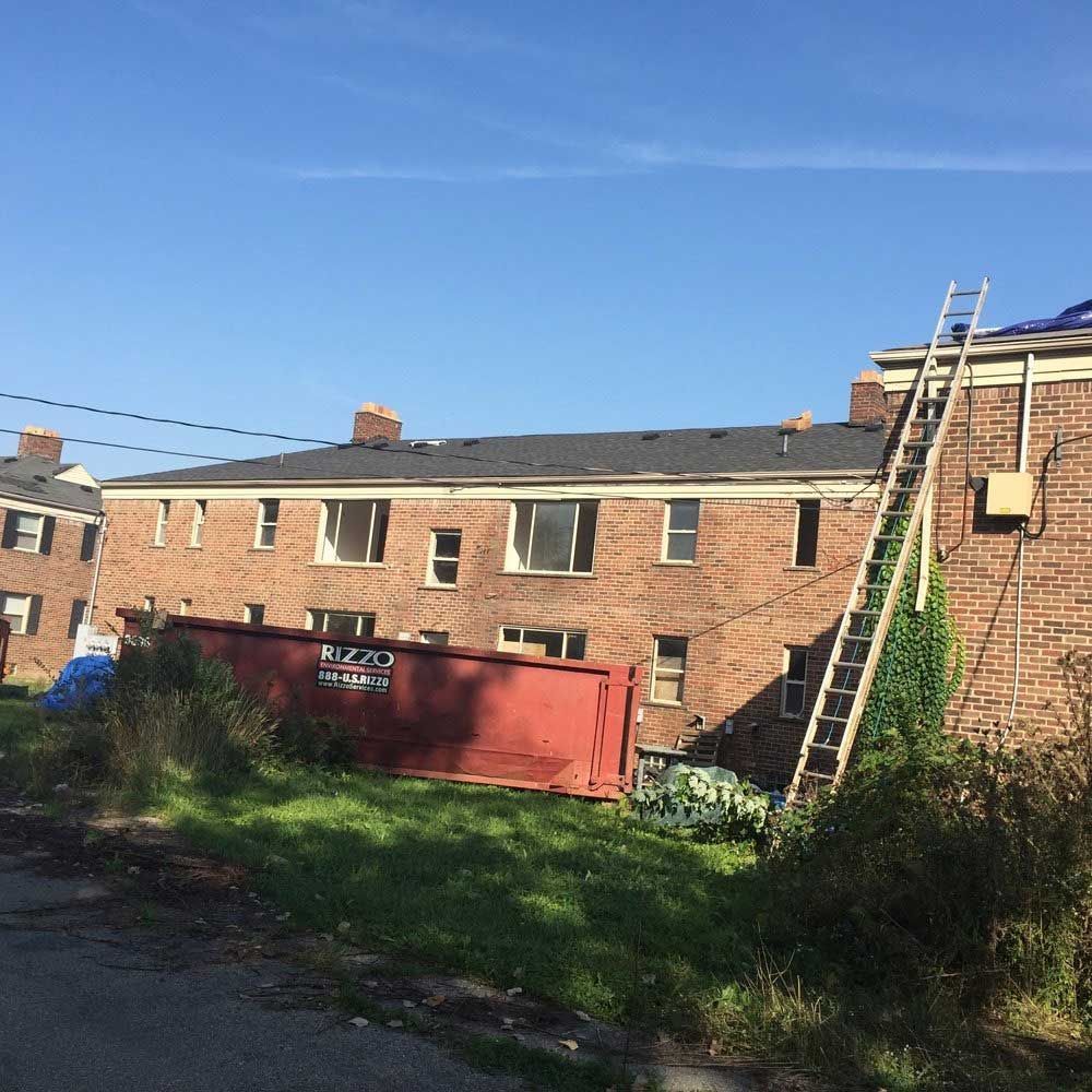 A multi-story brick apartment building under construction with a ladder leaning against it and a large red dumpster nearby.
