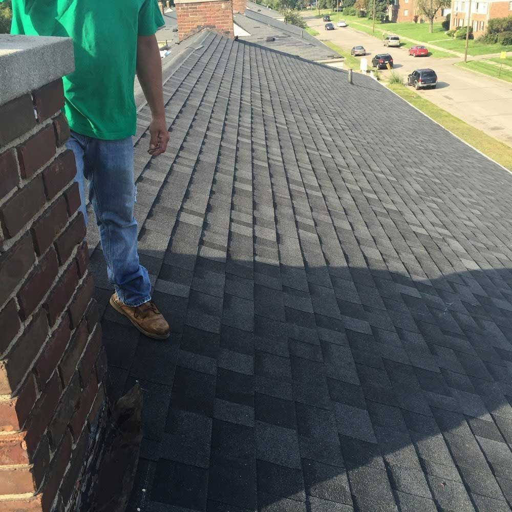 A person in a green shirt stands on a dark shingled roof next to a brick chimney, overlooking a residential street.