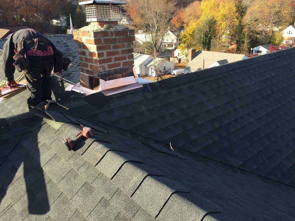 A worker in dark clothing installs shingles on a residential roof near a brick chimney on a sunny day.