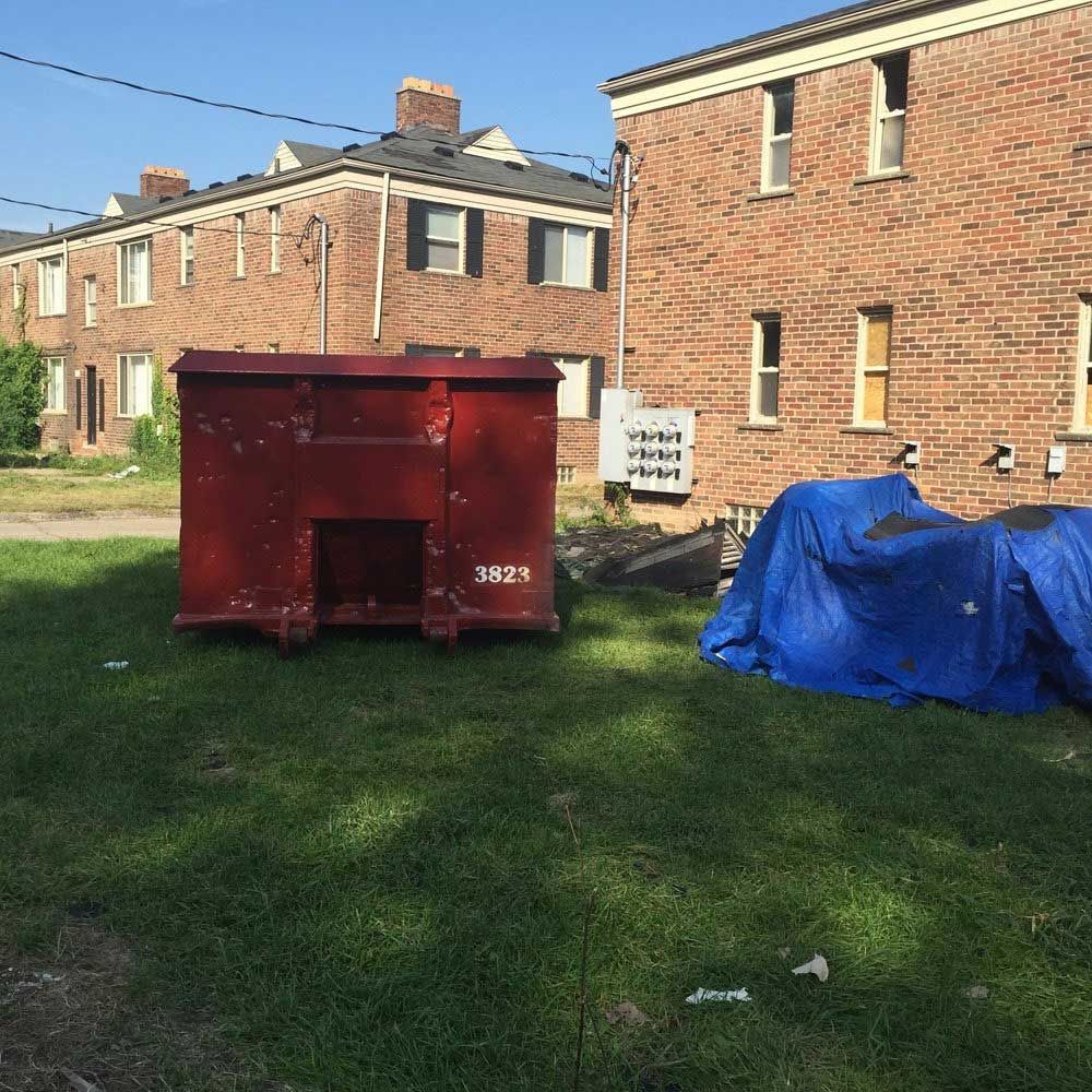 A large, dark red dumpster and a blue tarp covering equipment sit on a patch of grass in front of brick apartment buildings.