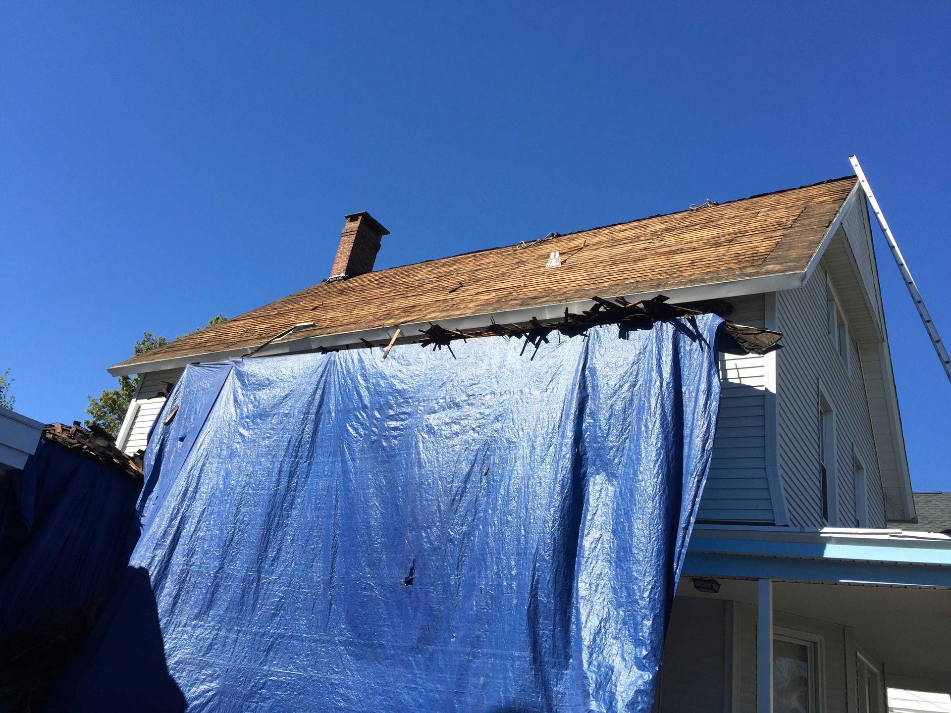 A house with a damaged roof partially covered by a large blue tarp against a clear blue sky.