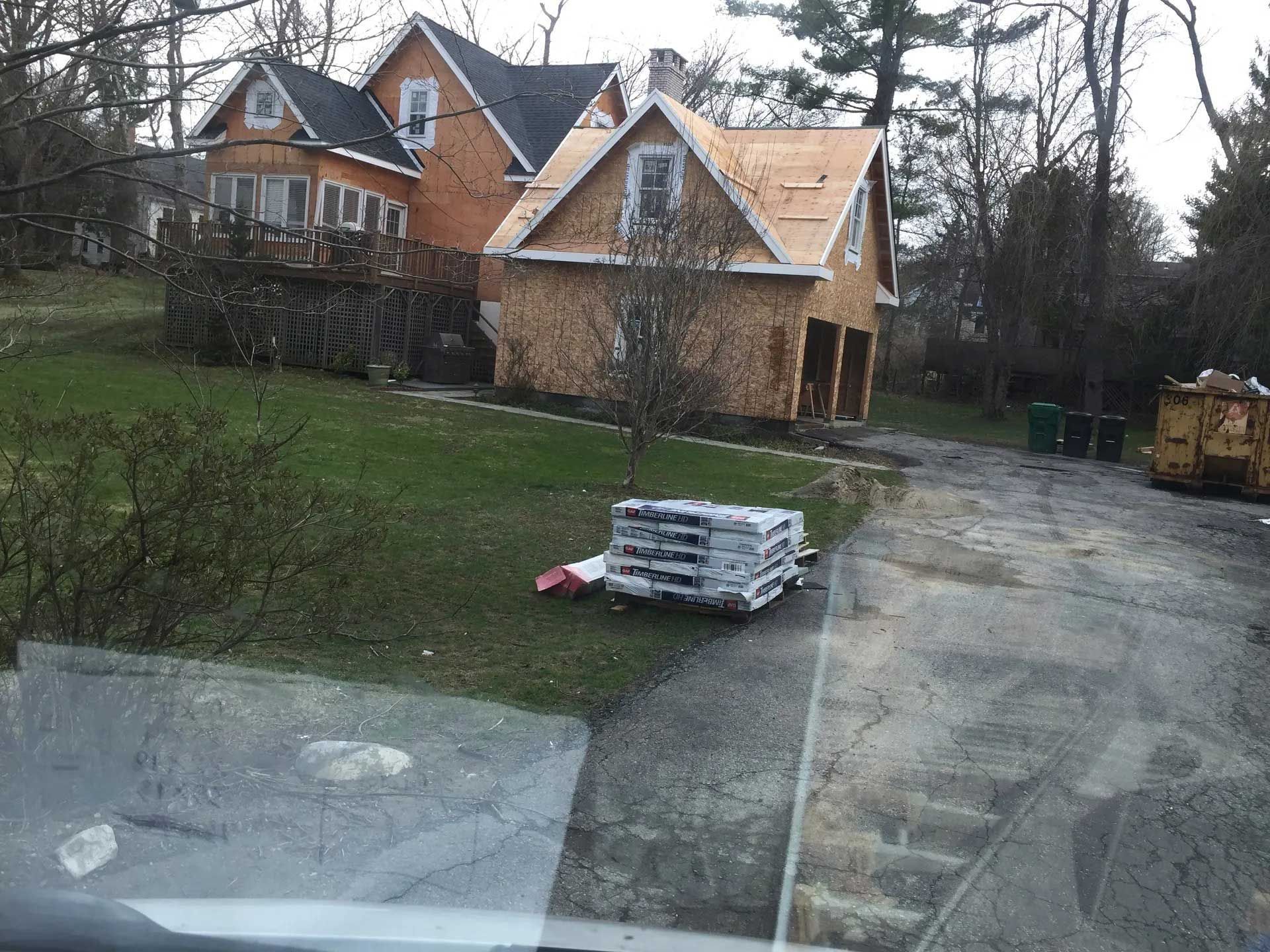 A construction site with a residential house and a new garage structure under construction, with materials on the lawn.