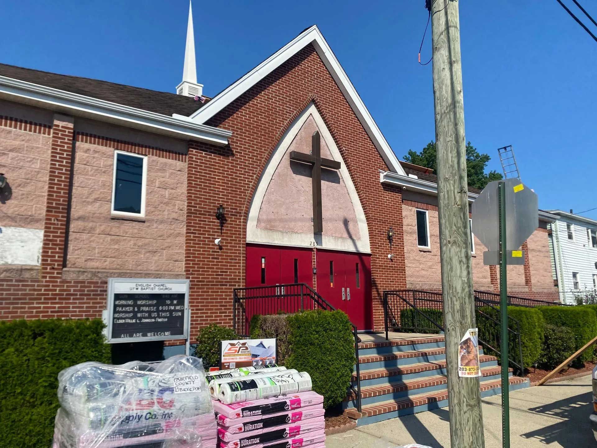 A brick church with a white steeple, a large cross above red double doors, and bags of mulch stacked on the sidewalk.
