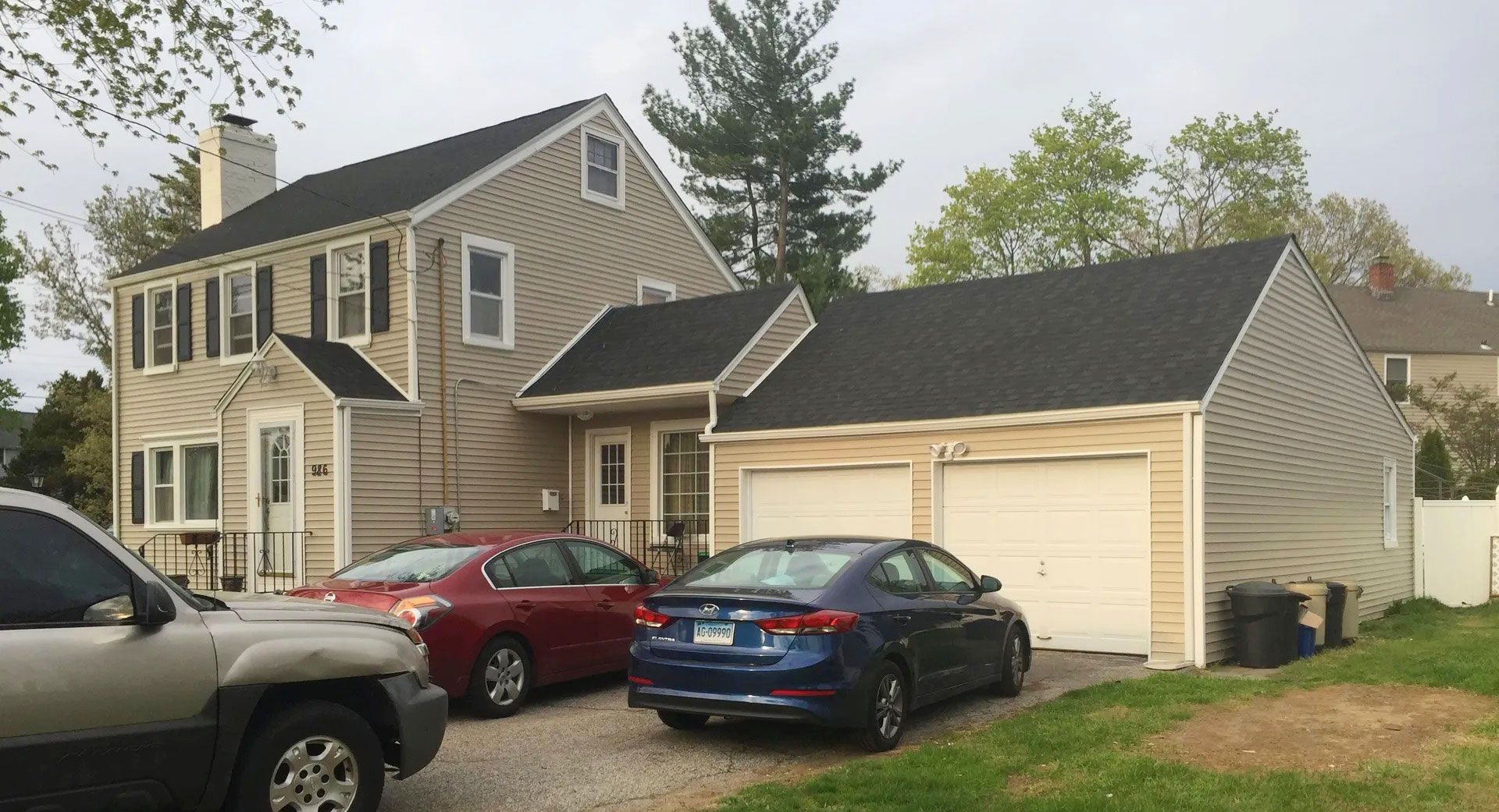 A tan, two-story house with an attached two-car garage, featuring a gravel driveway occupied by three parked cars.