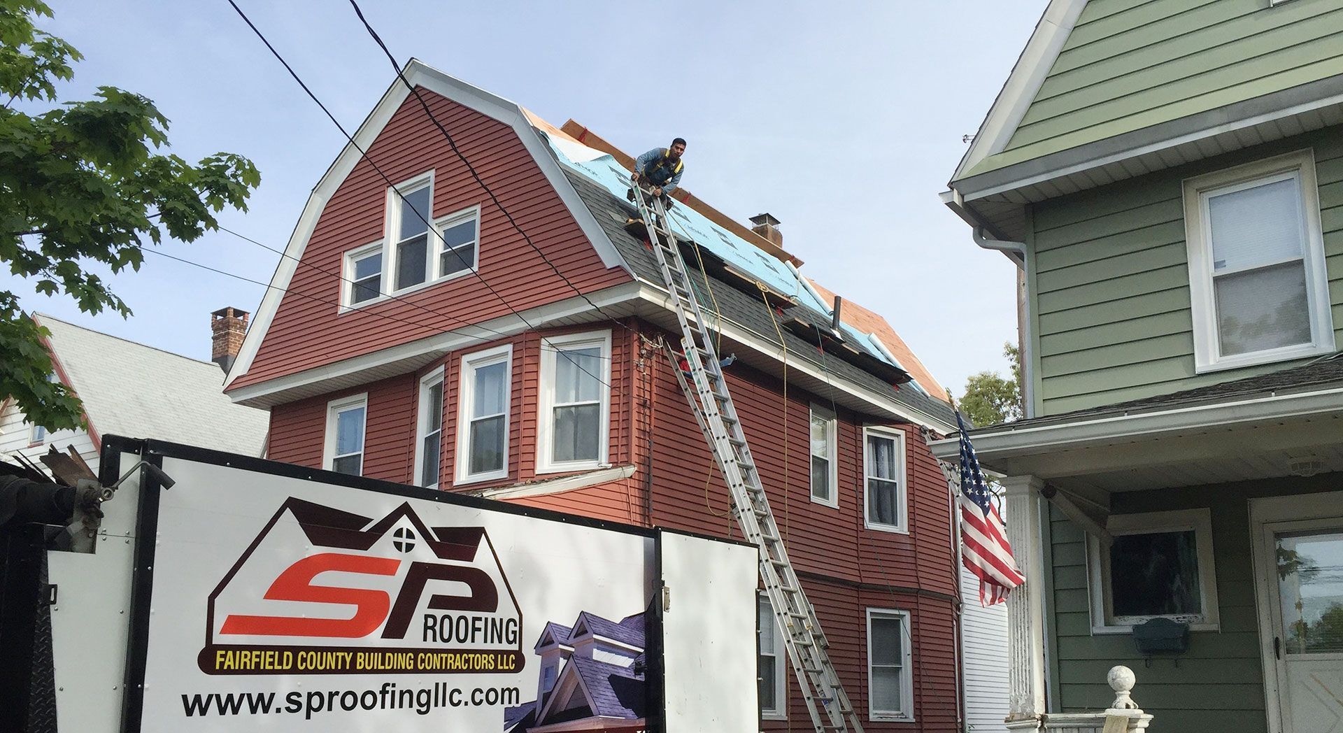 A construction worker on a roof in front of a red house, with an SP Roofing truck parked in the foreground.