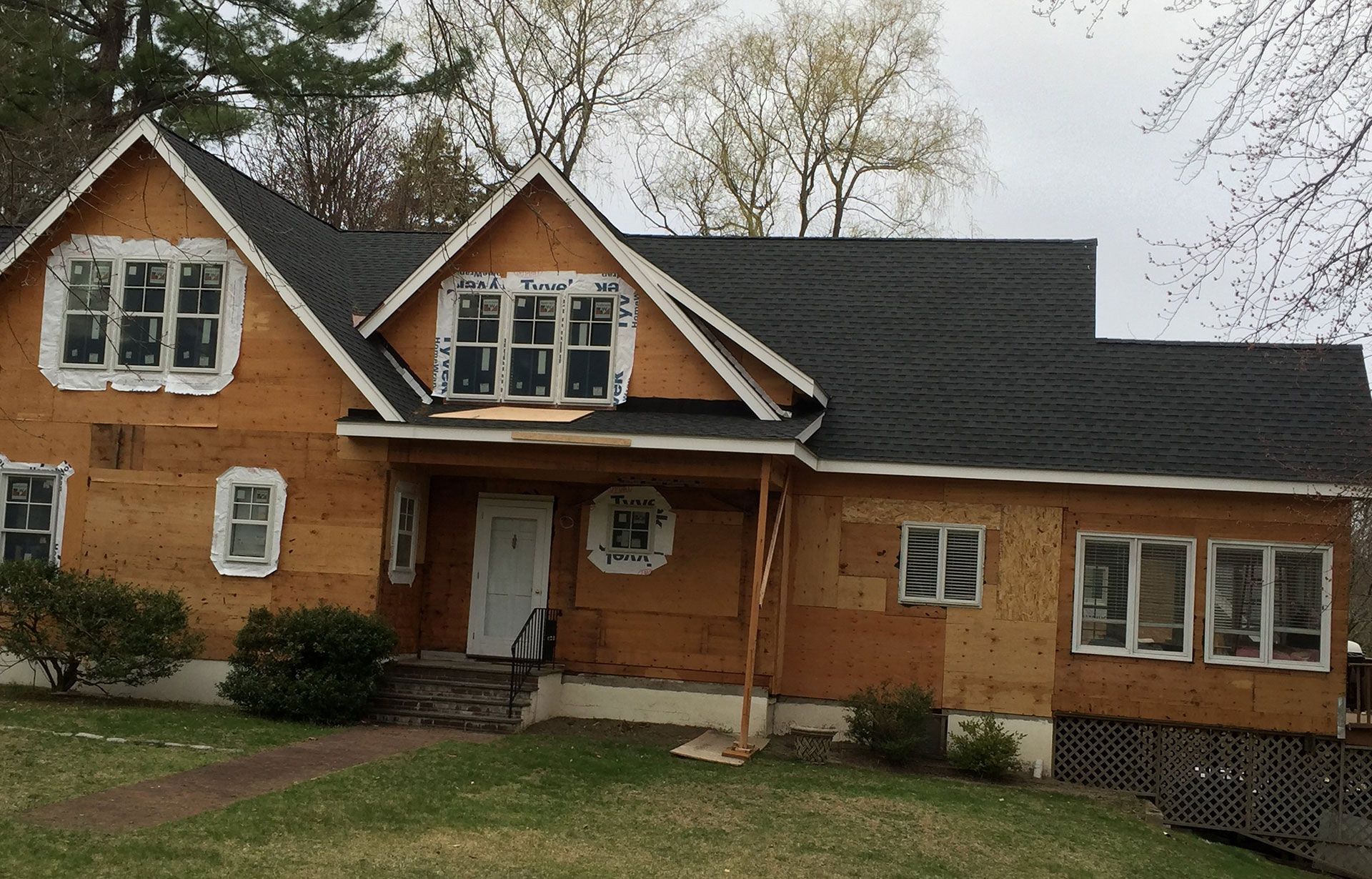 A two-story house under construction with exposed plywood siding and a newly installed black shingle roof.